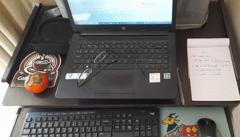 Image for Blogging - On a writing desk - a laptop is open with an external keyboard, mouse, and colourful mousepad with the image of a multi-coloured bull. To the right is a notepad with writing on it. To the left is an orange fruit, atop a coaster. A pair of reading glasses rests upon the laptop keypad.